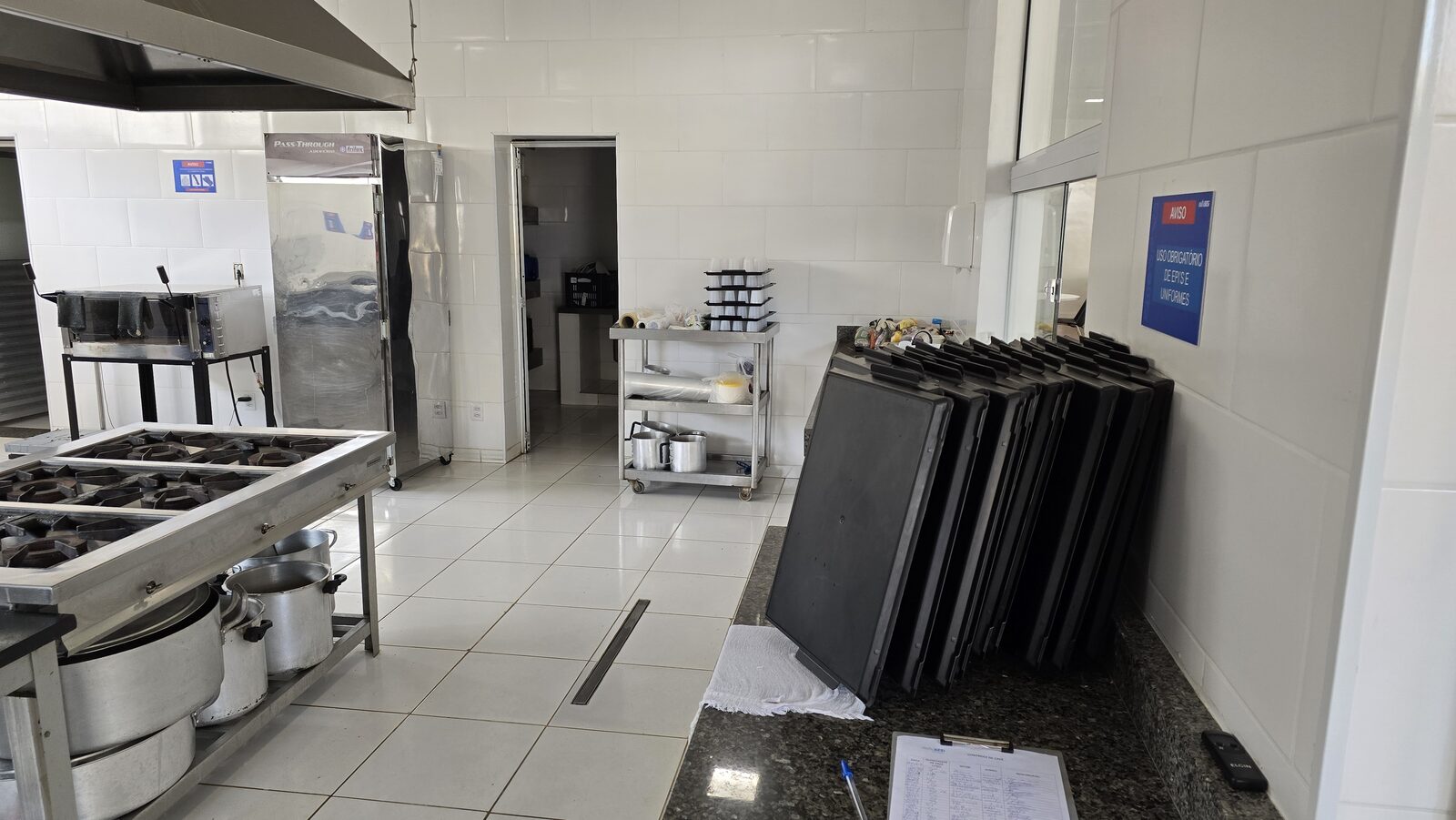 Esquadrias de vidro tipo pass-through entre a cozinha e o salão do refeitório, permitindo controle visual e comunicação entre os ambientes